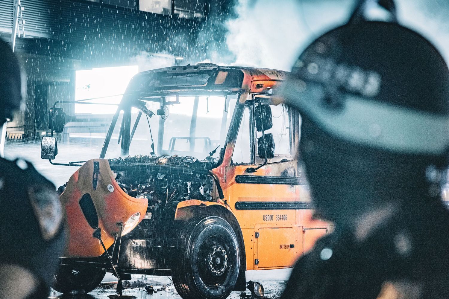 Firefighters battling a burning orange forklift truck in a smoky industrial setting during an emergency response.