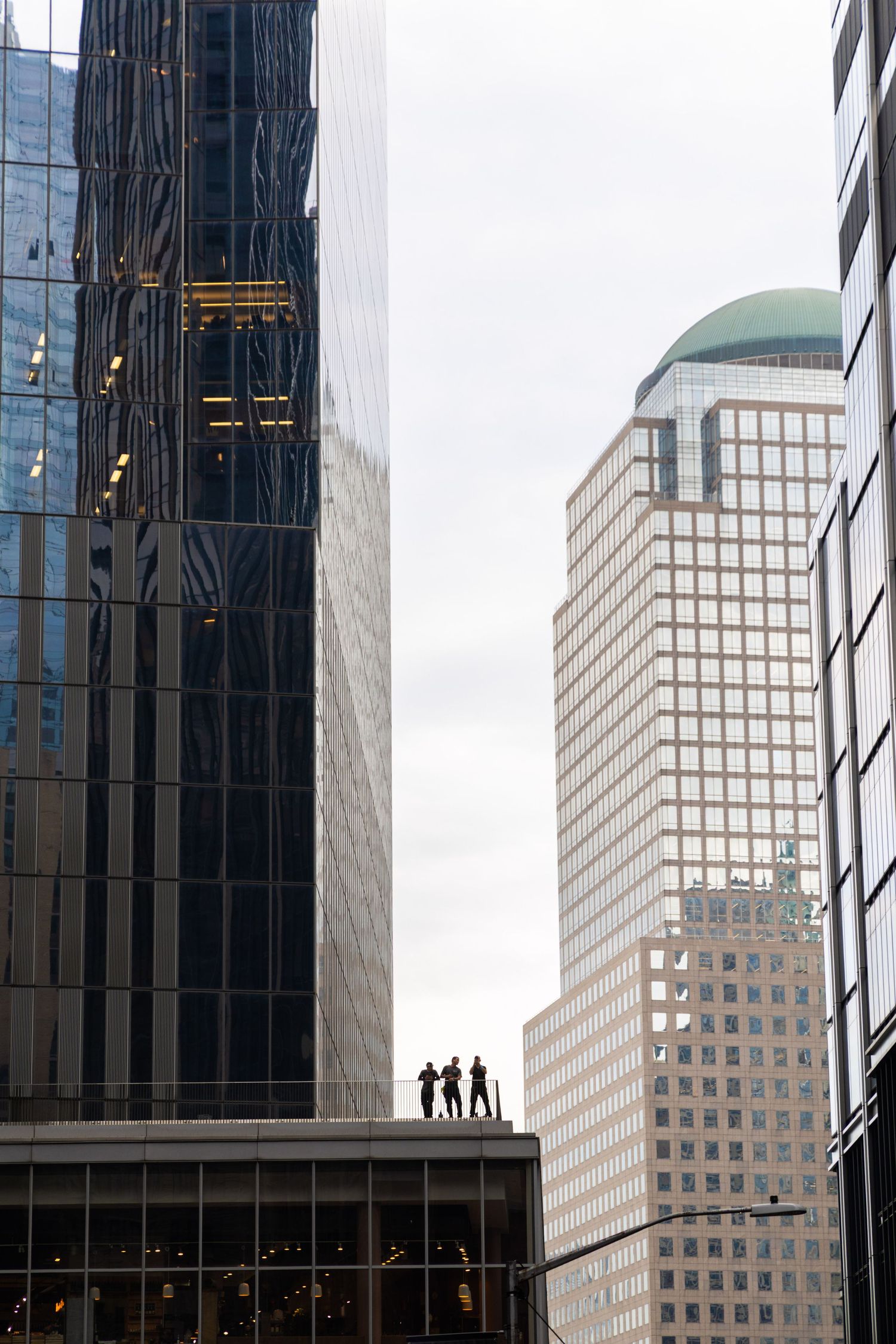 Three silhouetted figures stand on a rooftop surrounded by towering glass and stone skyscrapers in an urban cityscape.