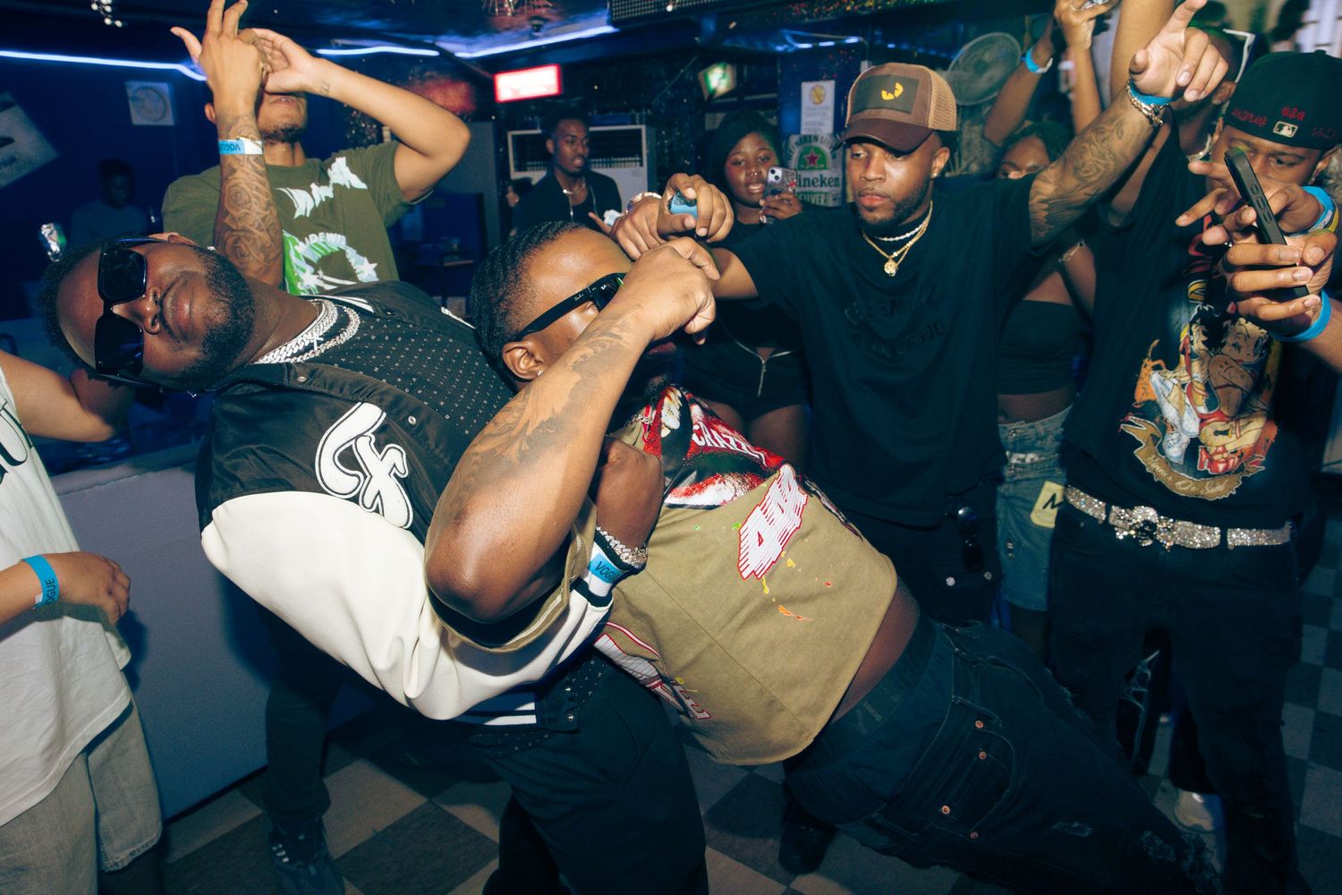 People dancing energetically at a nightclub party, with crowd gathered around in a dimly lit venue.