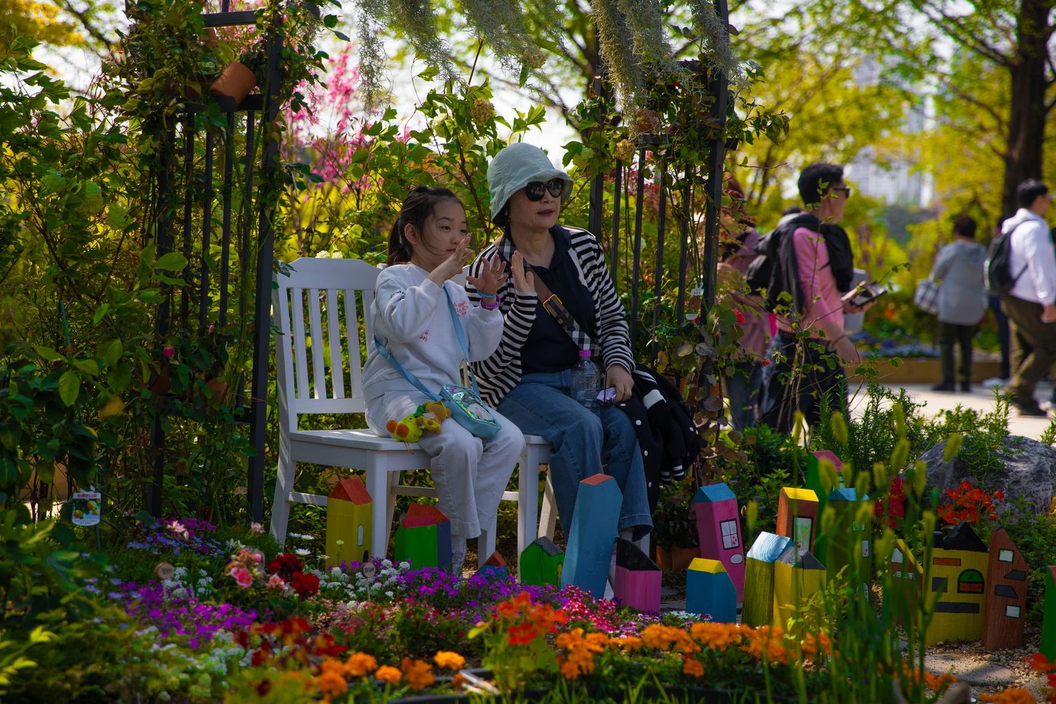 Woman and child sitting on a white bench surrounded by colorful flowers in a garden display.