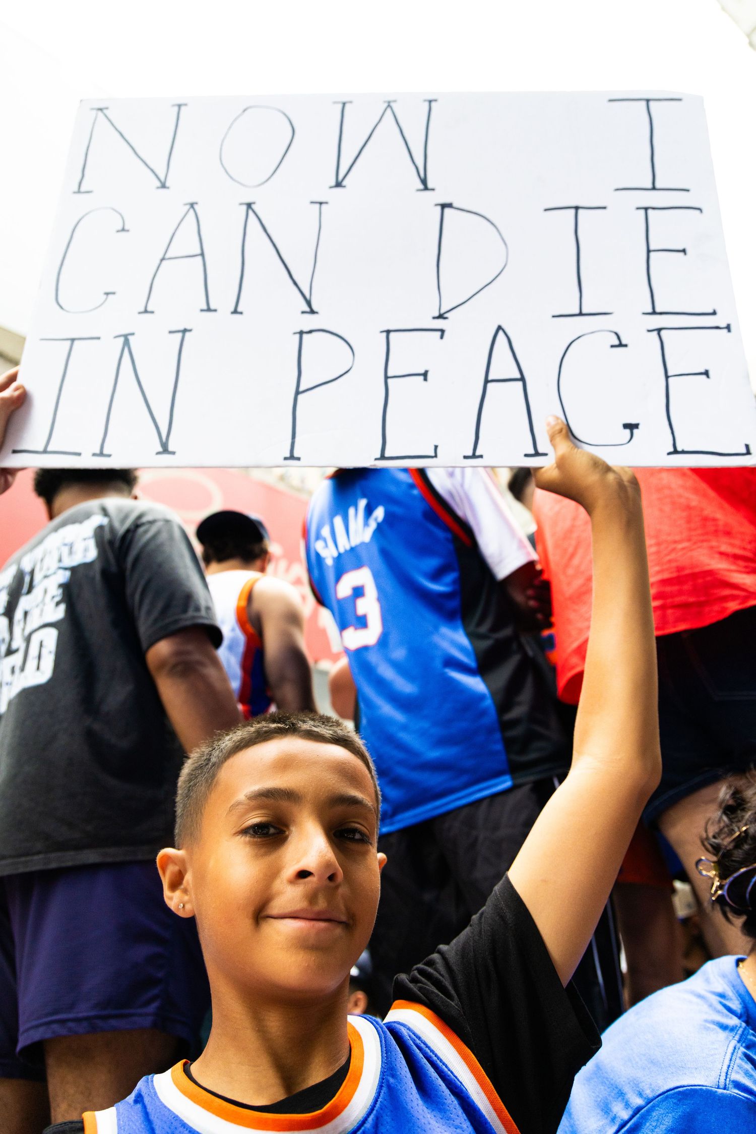 Young boy at a sports celebration holding a sign reading Now I Can Die In Peace among a crowd of fans.