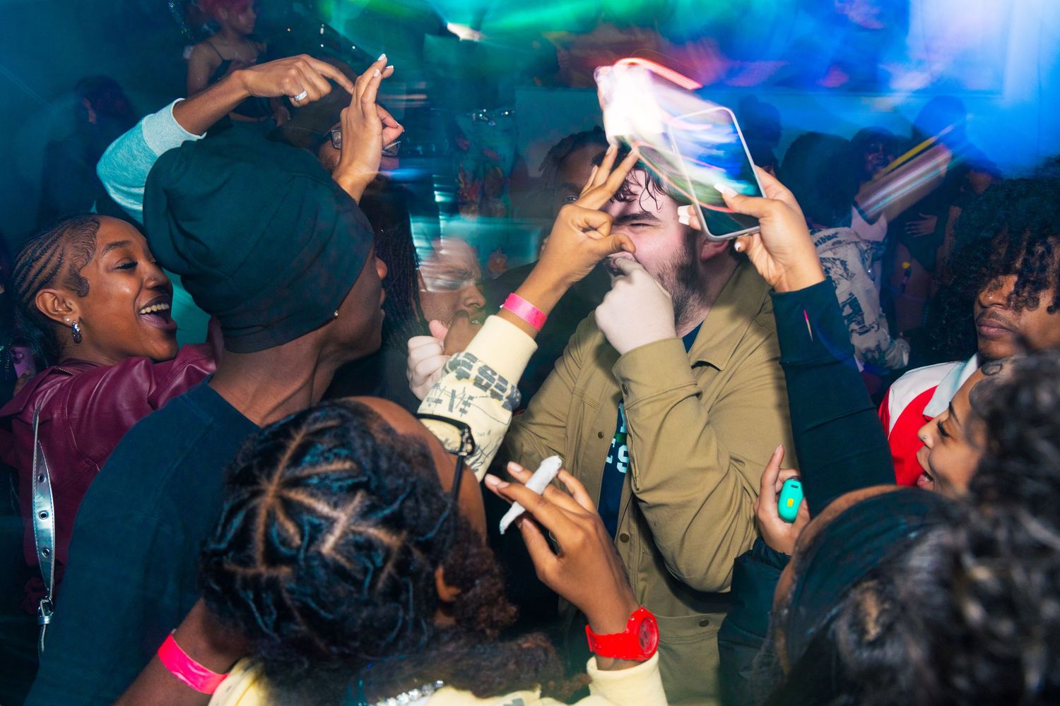 Crowd of people dancing and taking selfies at a lively nightclub party with colorful lights.