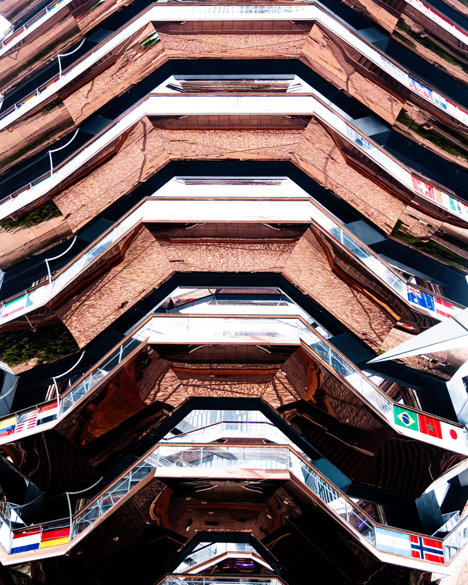 Looking up at The Vessel structure in Hudson Yards NYC, featuring honeycomb staircases adorned with international flags.