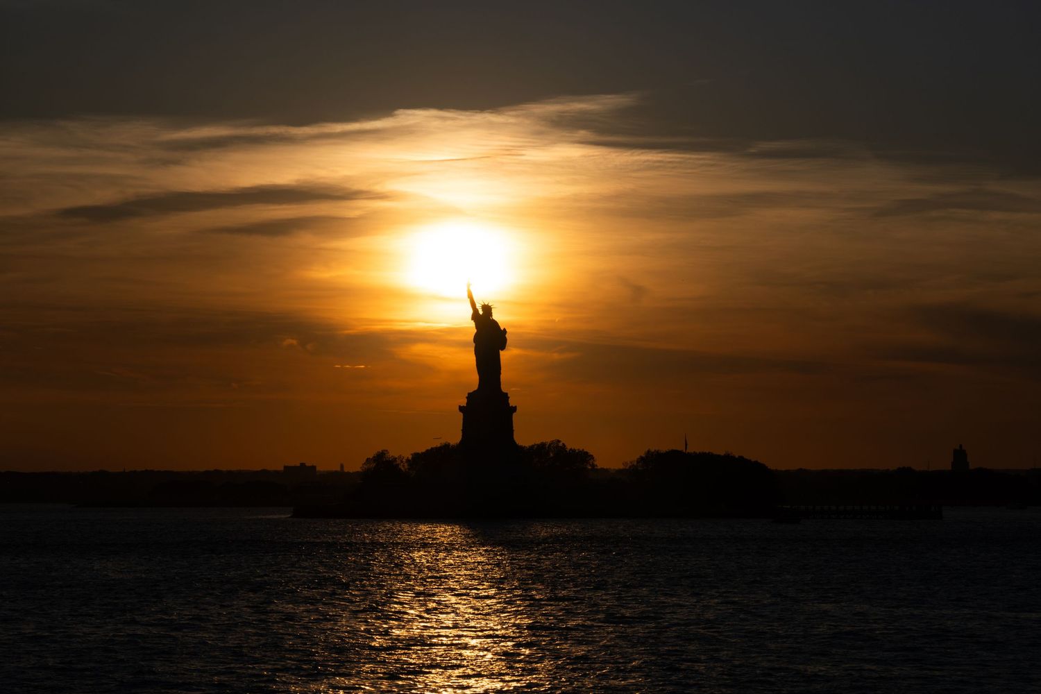 Silhouette of the Statue of Liberty at sunset with golden sky reflecting on calm water in New York Harbor.