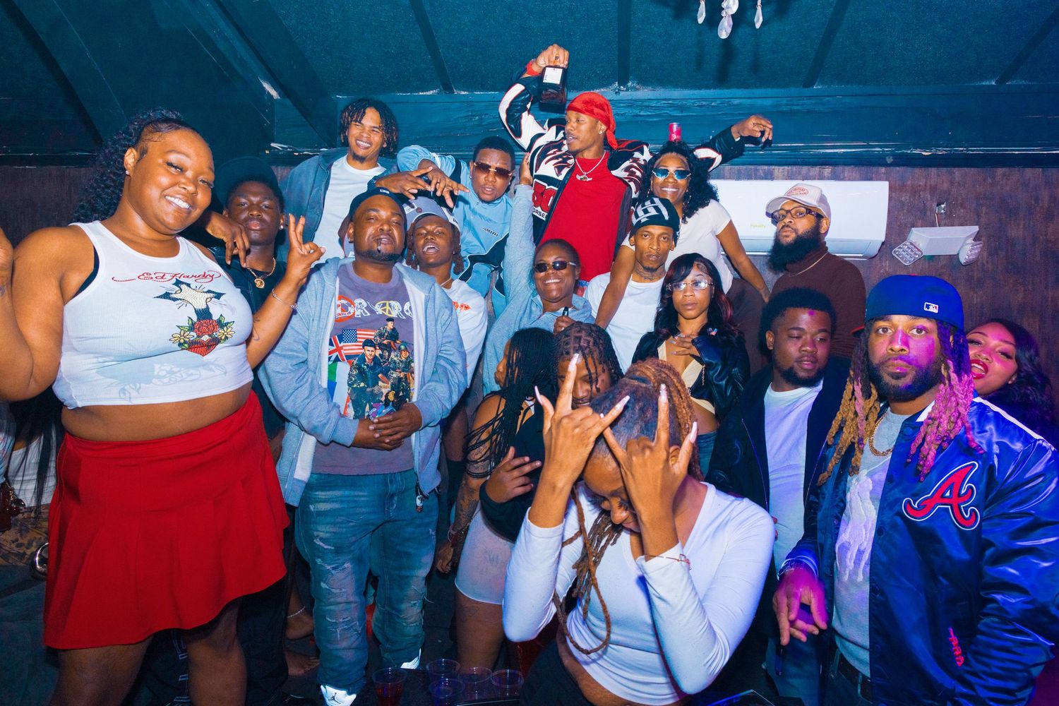 Large group of friends posing together at an indoor party, smiling and making hand gestures under dim lighting.