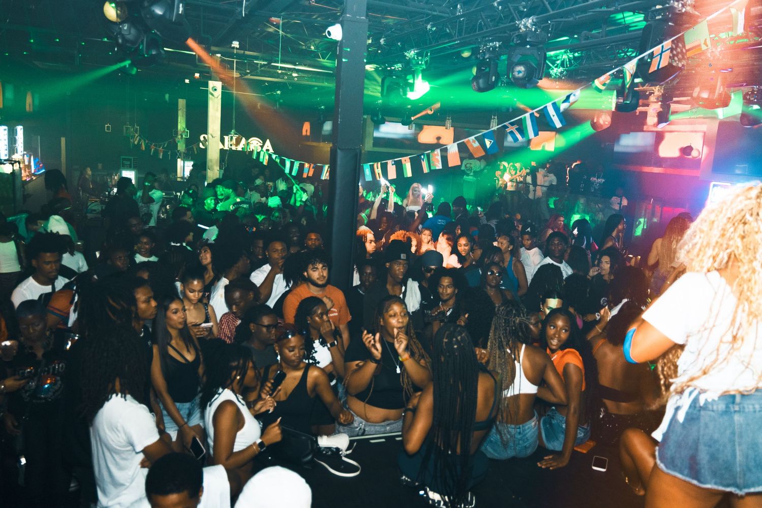 Energetic nightclub crowd dancing under green lights with colorful bunting decorations at a packed party event.