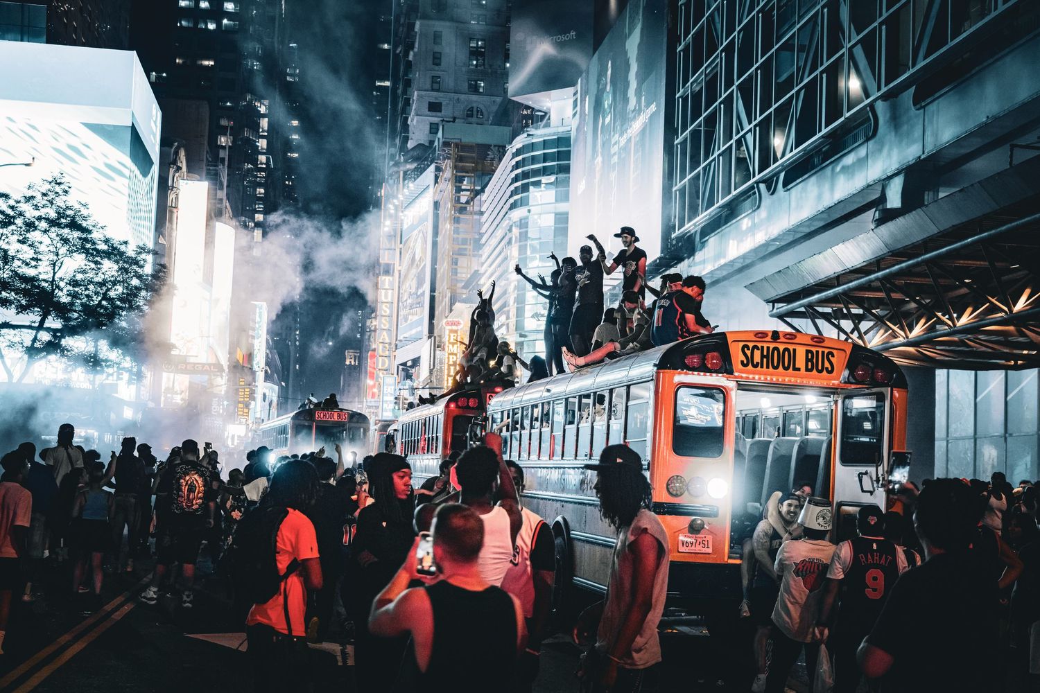 Crowd of people surrounding a school bus on a city street at night with smoke and tall buildings.