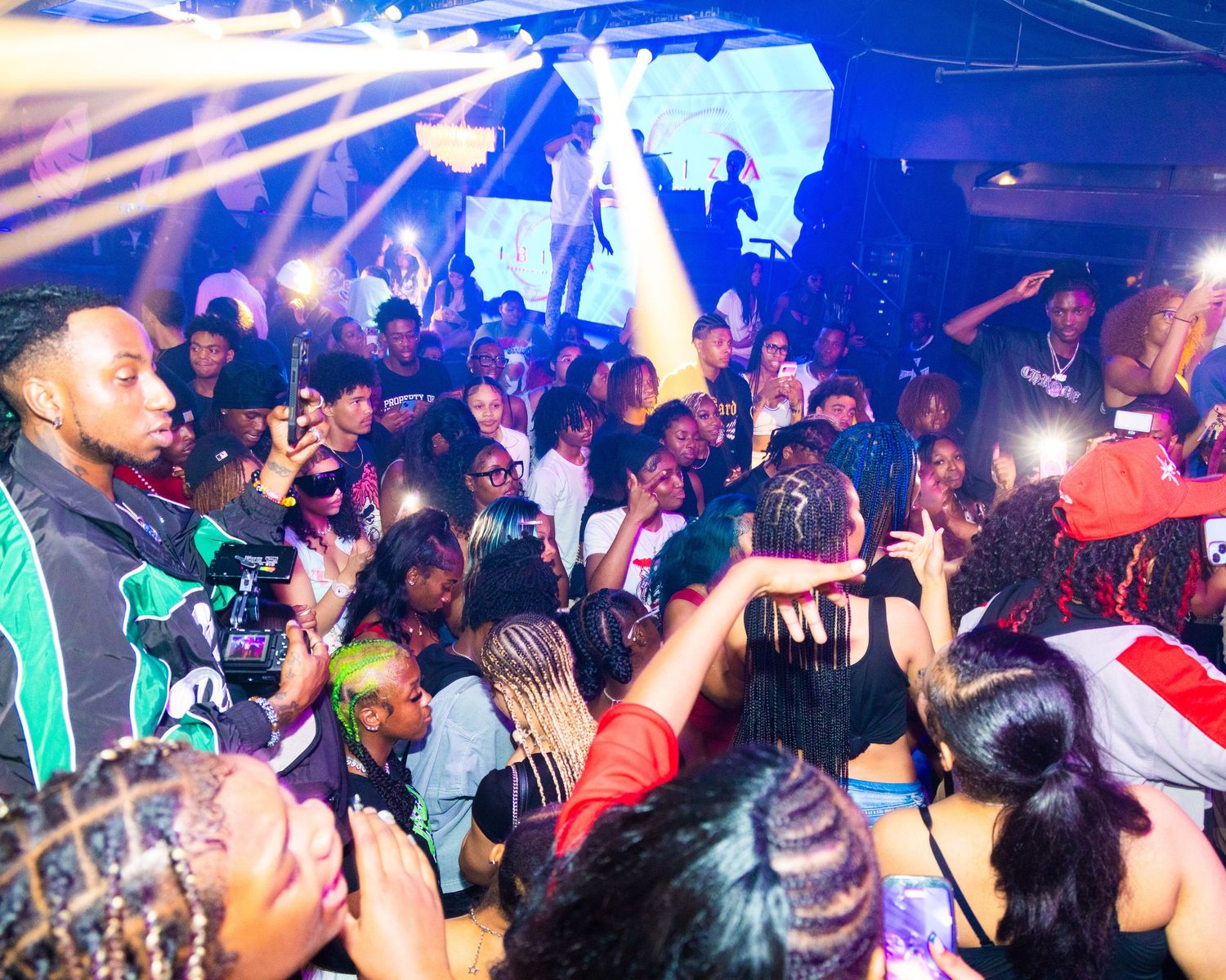Energetic nightclub party scene with a large crowd dancing under blue lights, people raising phones to capture the moment.