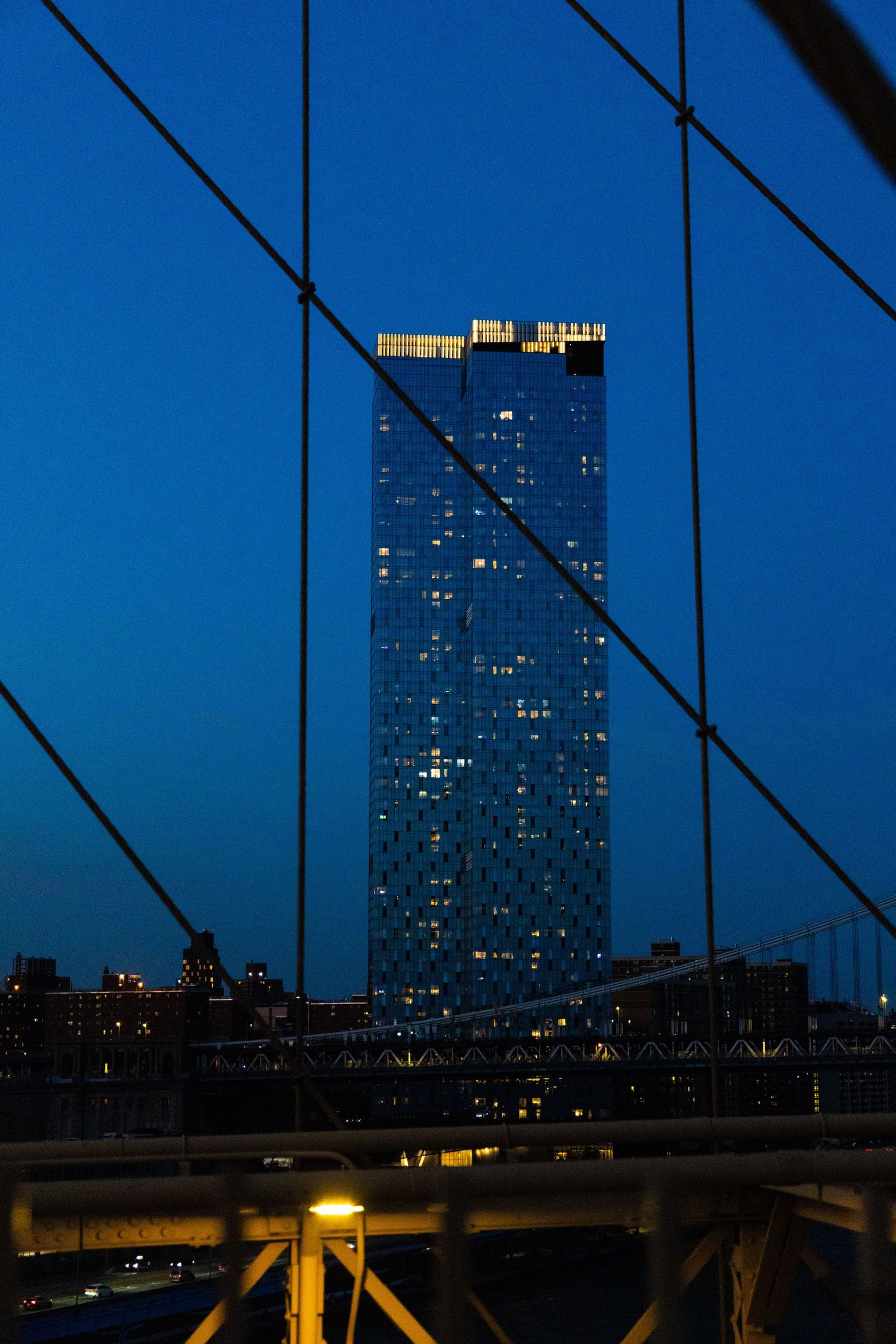 Night view of a glass skyscraper through bridge cables and steel girders against a deep blue twilight sky.