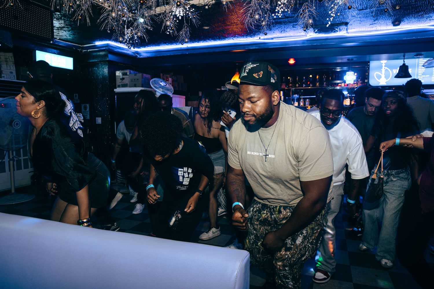 Man in beige shirt stands confidently near a bar in a busy nightclub setting.