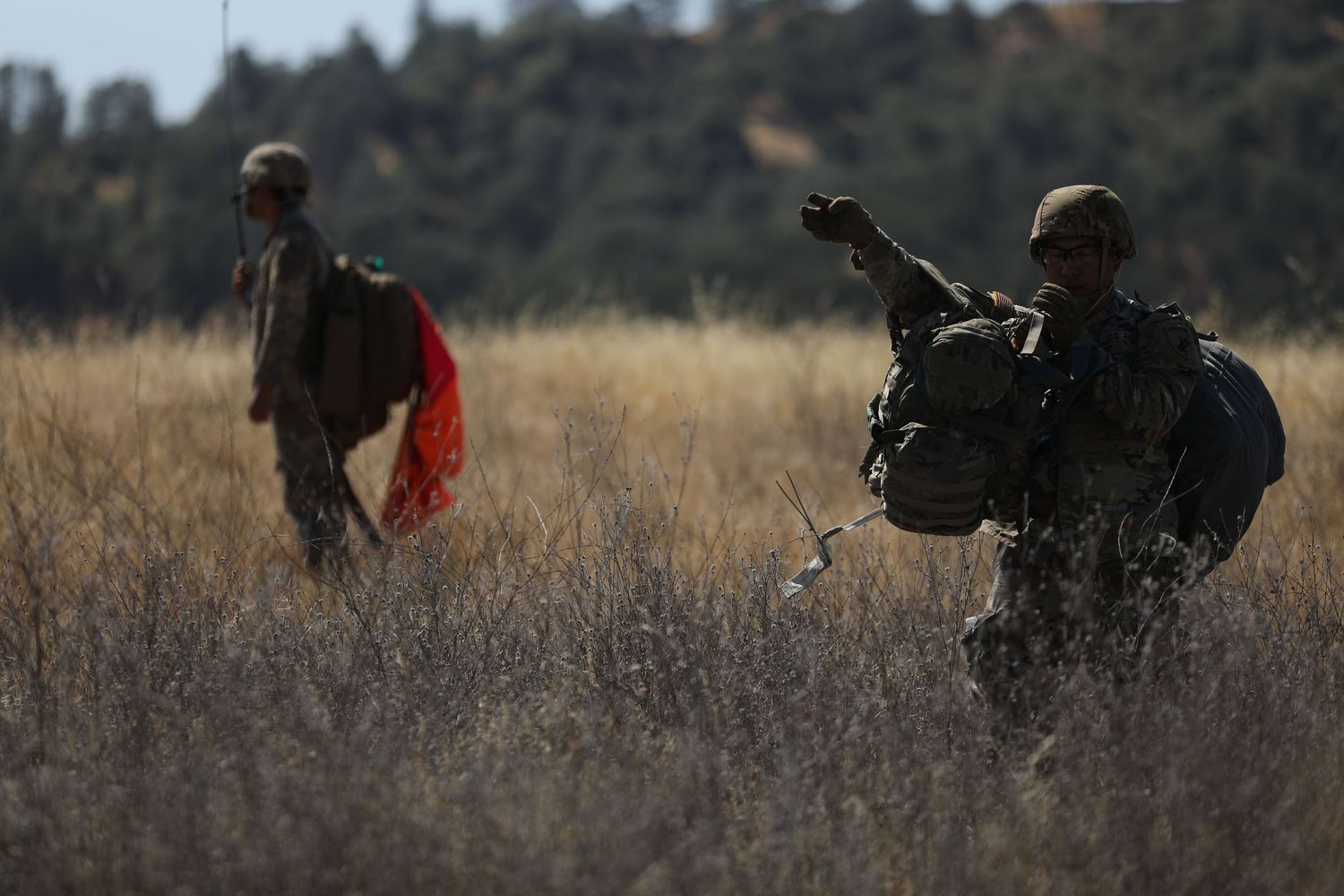 Two soldiers in full combat gear walk through a dry grass field carrying equipment after an airborne operation.