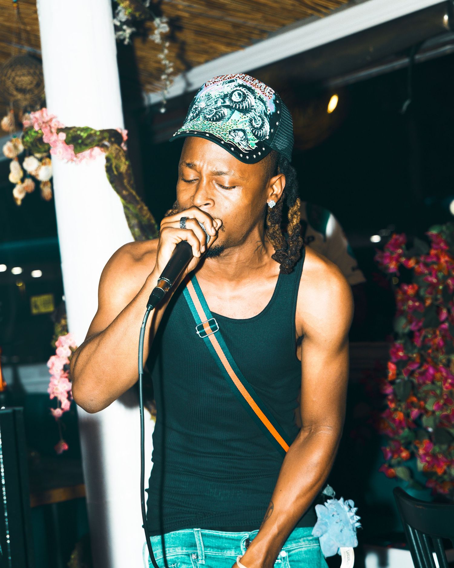 A male performer sings passionately into a microphone at an outdoor venue, wearing a black tank top and patterned cap.