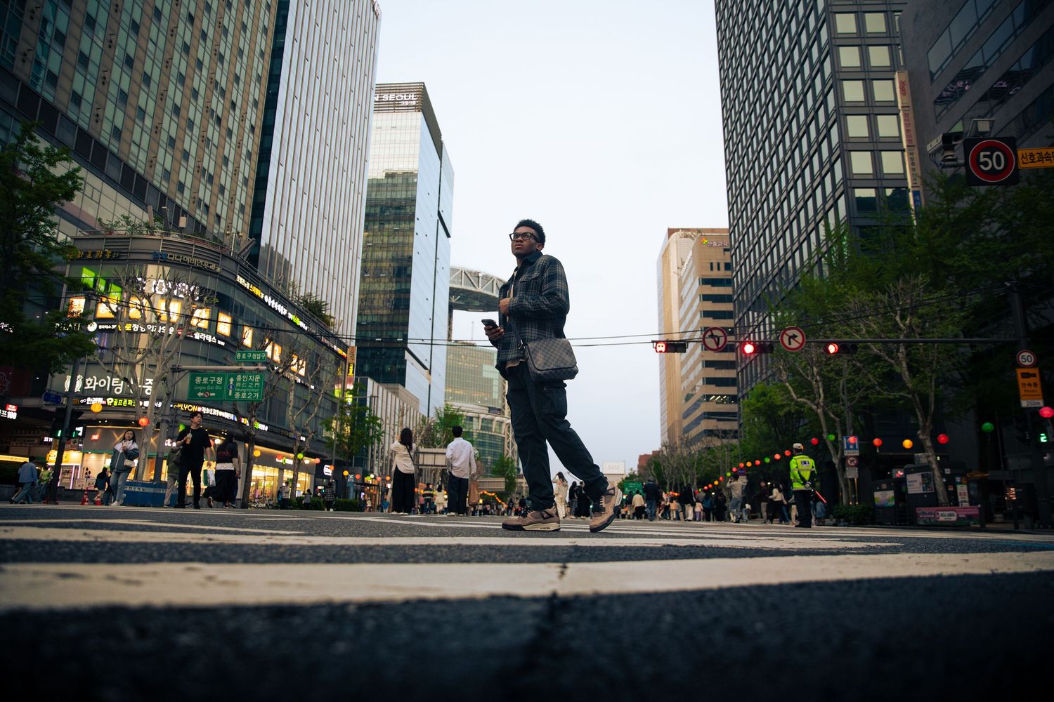 Person crossing a busy urban intersection surrounded by modern skyscrapers and green trees at dusk.