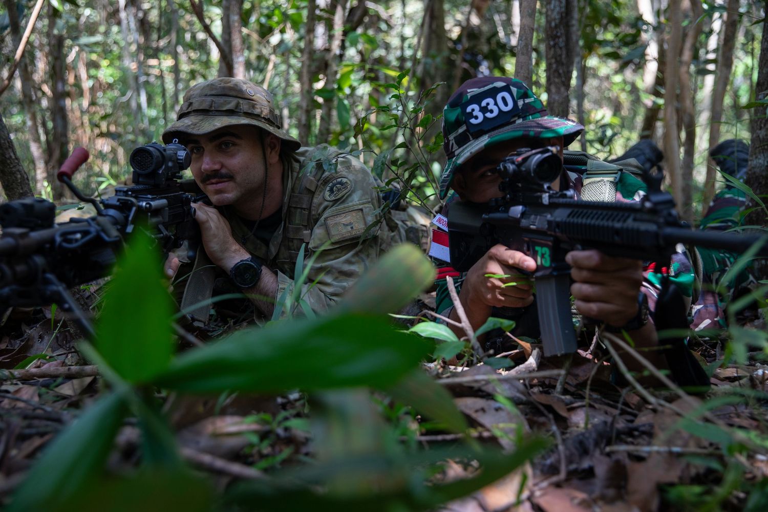 Two multinational soldiers aim rifles from a prone position in dense jungle undergrowth during combat training.