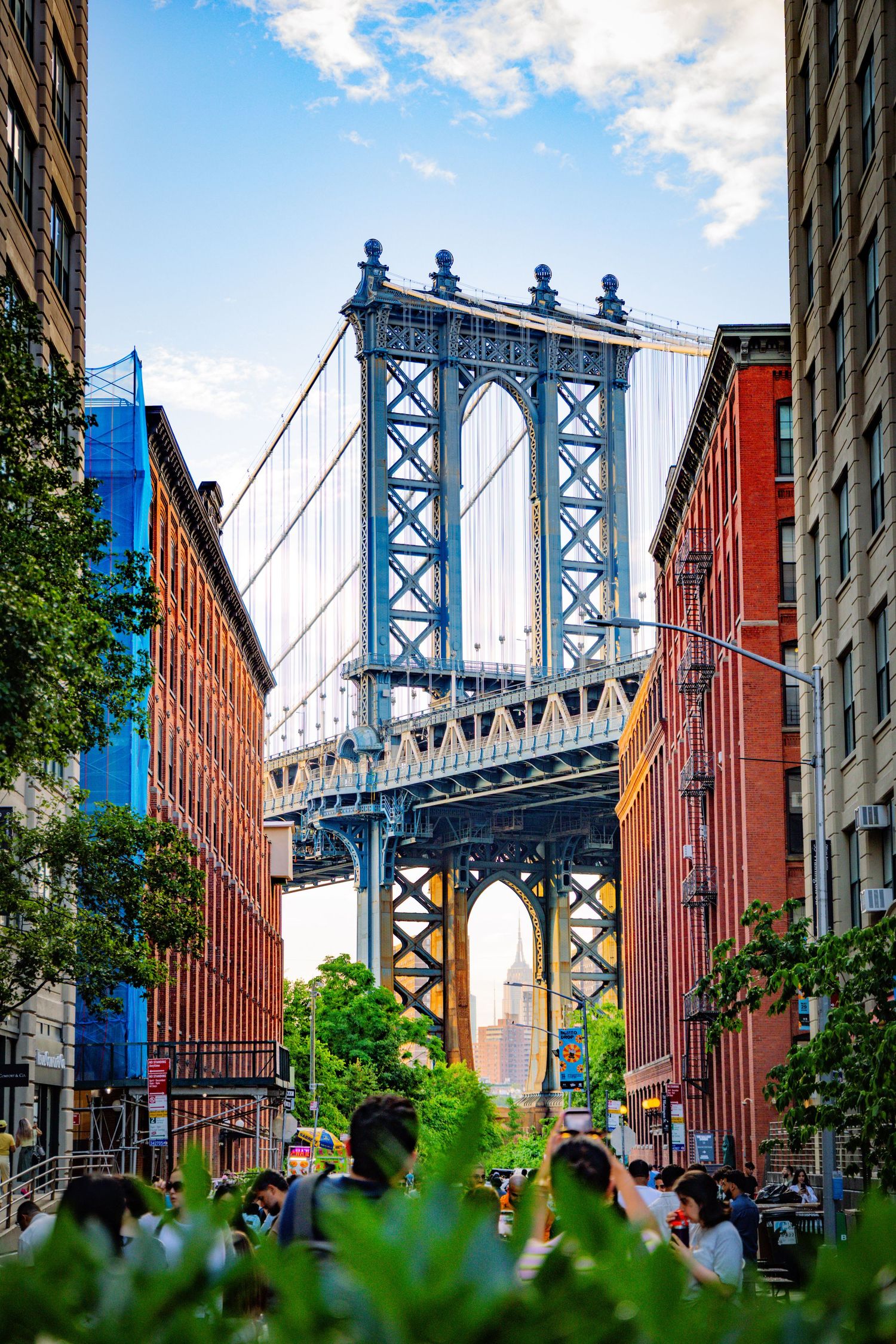 Manhattan Bridge tower framed by DUMBO Brooklyn brick buildings with crowds below and blue sky backdrop.