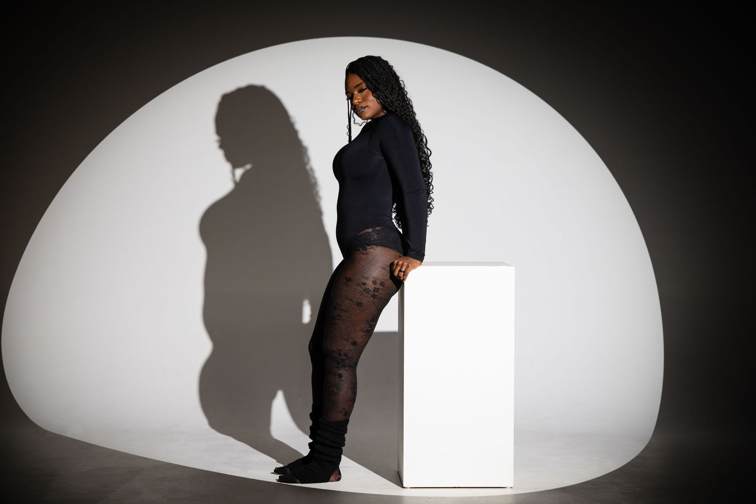 Woman in black outfit leaning on white pedestal with dramatic shadow on curved white background.