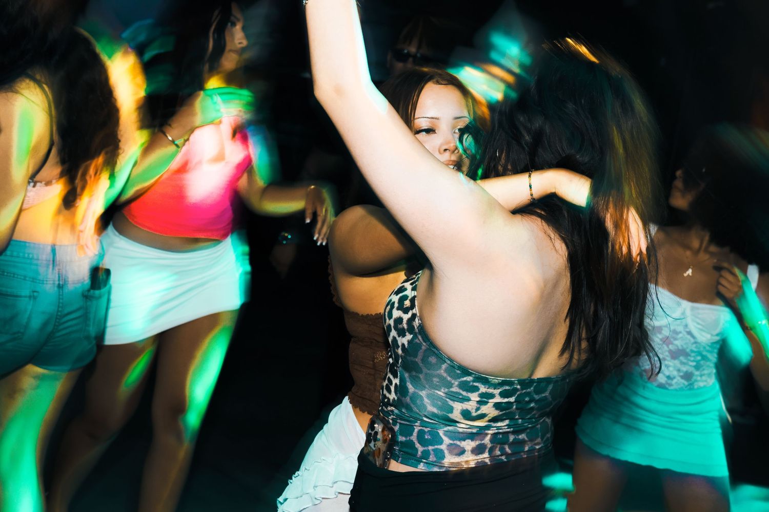 Young women dancing energetically at a nightclub with colorful green and pink lighting.