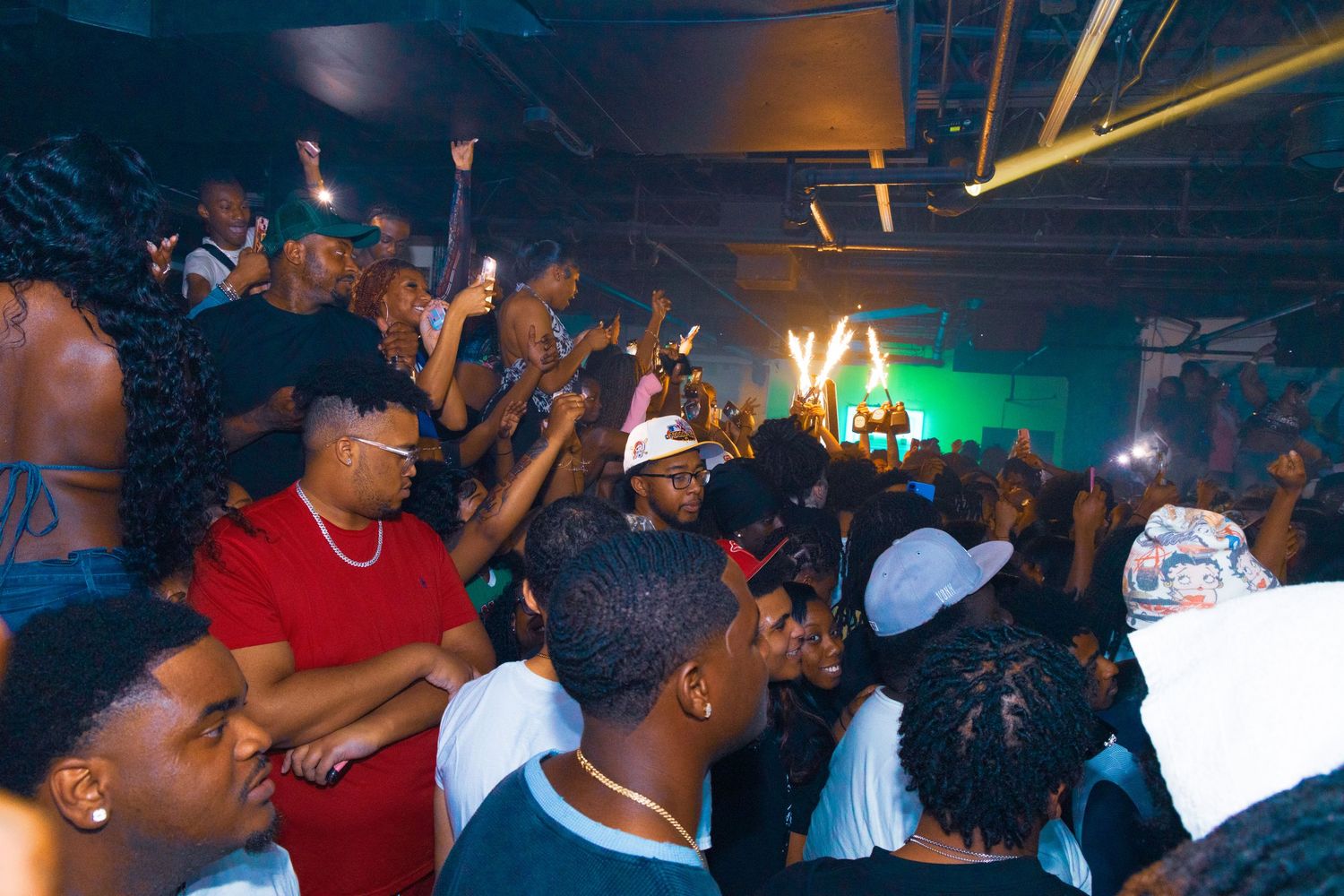 Crowded nightclub party with people dancing and raising hands under colorful lights in a dark venue.