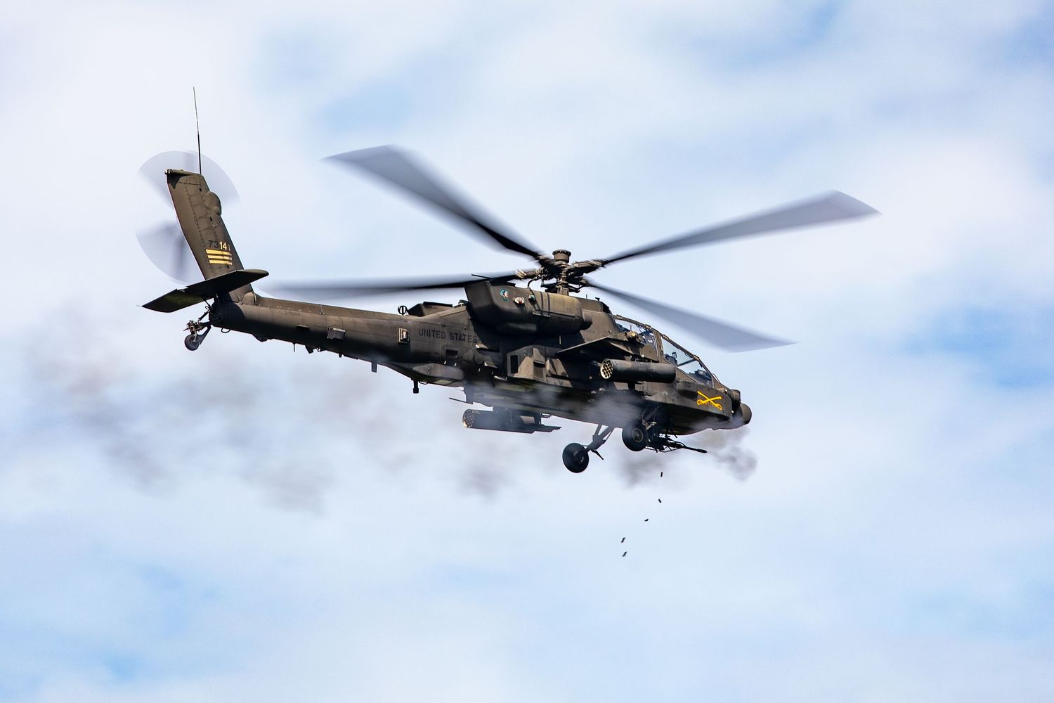 Military Apache attack helicopter firing weapons amid smoke against a cloudy sky.