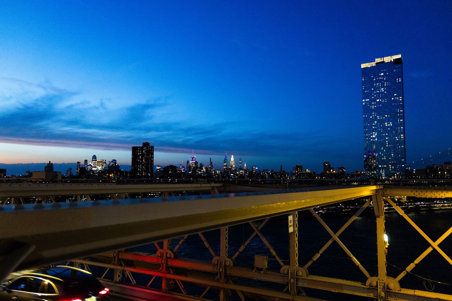 Night cityscape view from a bridge with glowing skyscrapers reflecting on water under a deep blue twilight sky.