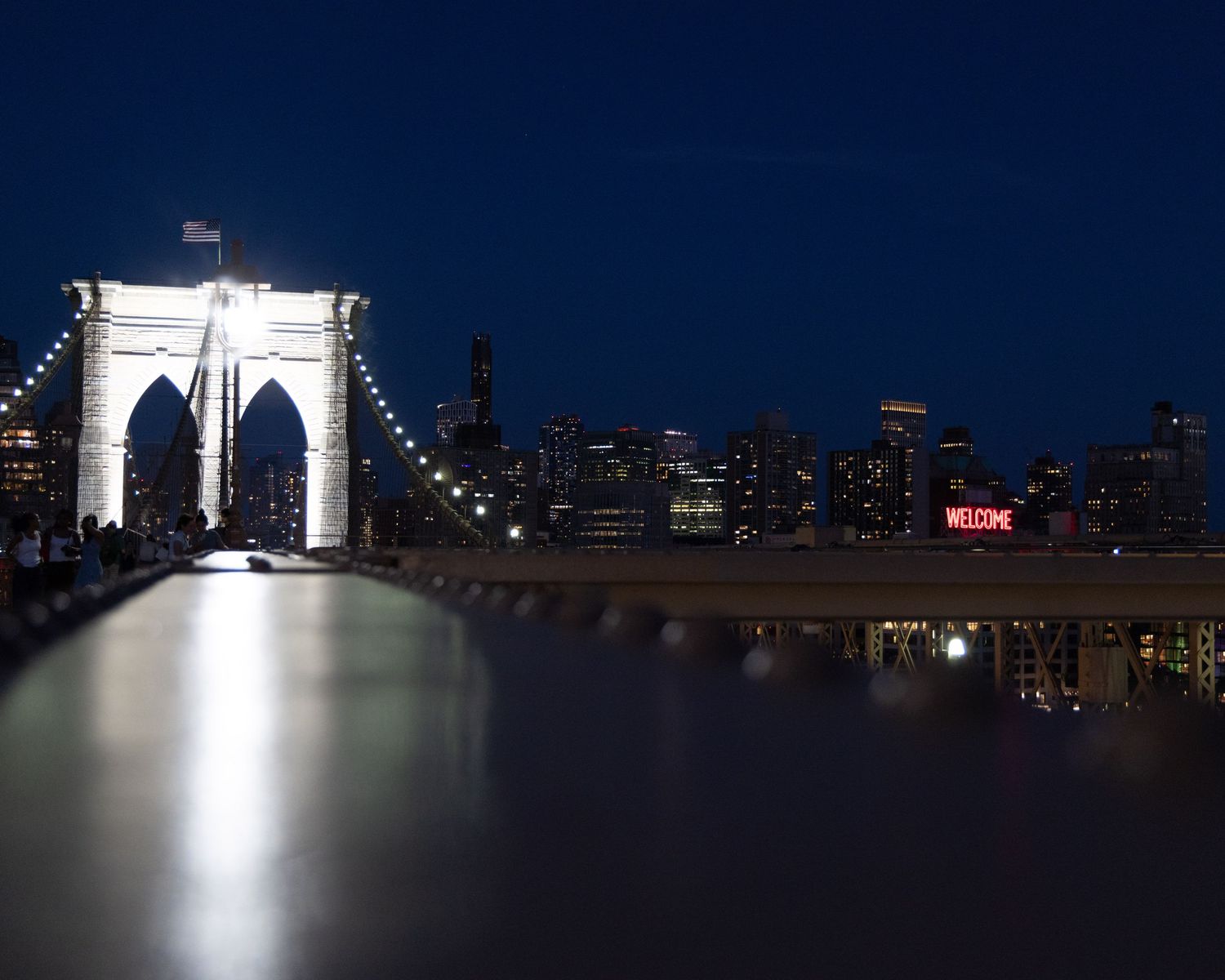 Brooklyn Bridge illuminated at night with NYC skyline reflecting on calm water below.
