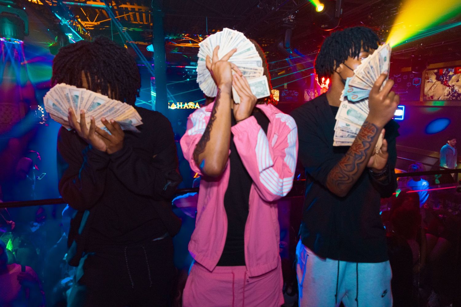 Three people in a nightclub holding fanned cash over their faces, with colorful neon lights in the background.