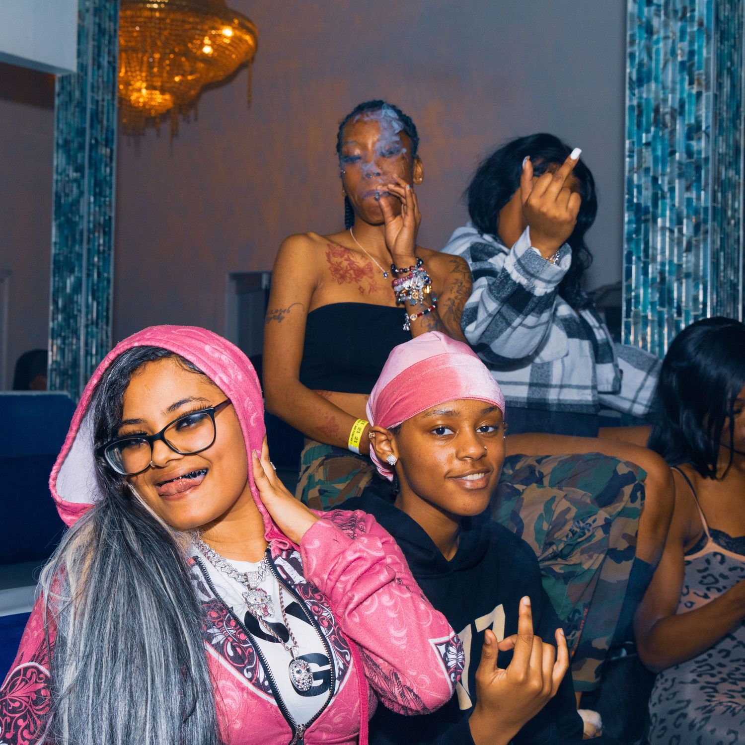 Group of young women posing together at an indoor party, wearing colorful outfits including pink hoodie and black top.