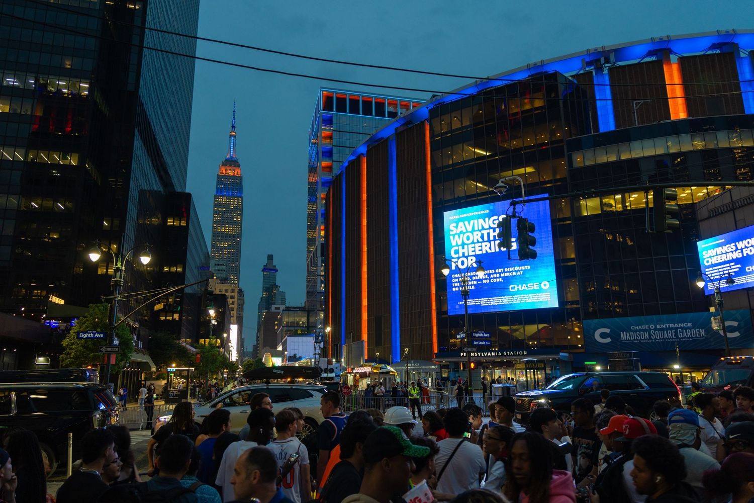 Crowded New York City street at night with illuminated Madison Square Garden and One World Trade Center skyline.