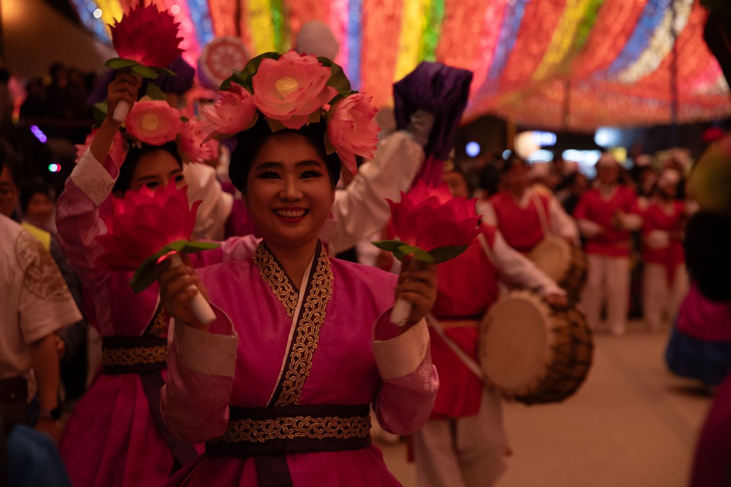 Smiling performer in pink traditional Korean costume holding lotus flowers at a night parade.