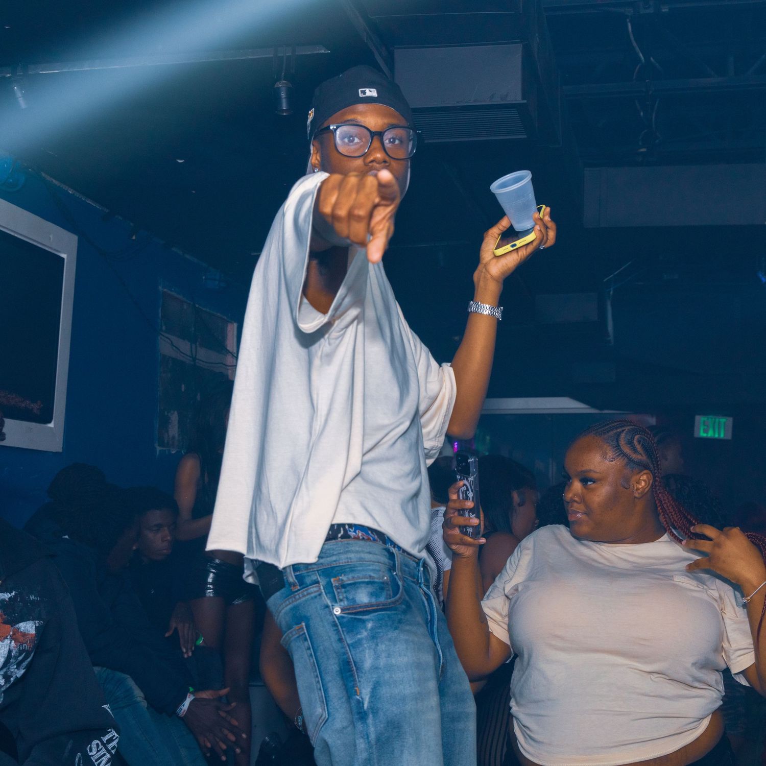 Man in white shirt and cap holds cup at a lively nightclub party with people dancing in background.