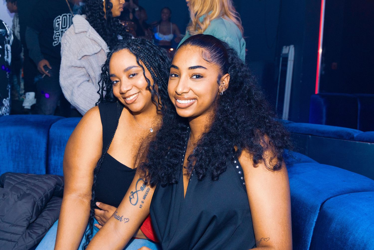 Two young women smiling together at a nightclub, sitting on a blue couch in dim blue lighting.