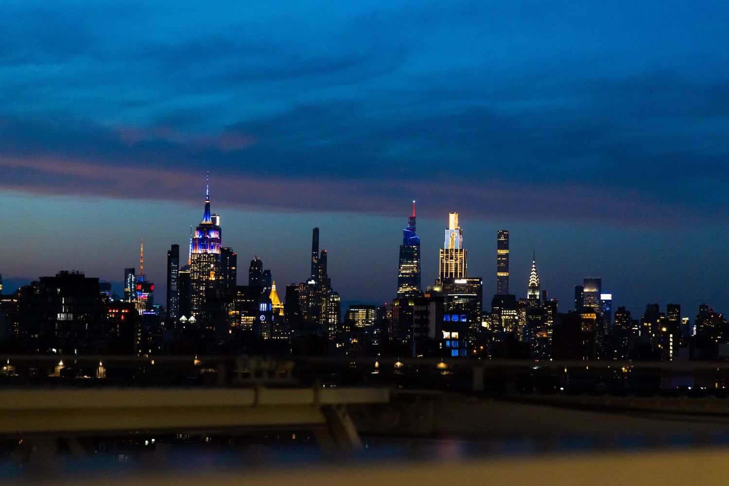 New York City skyline illuminated at dusk with colorful skyscrapers against a deep blue twilight sky.
