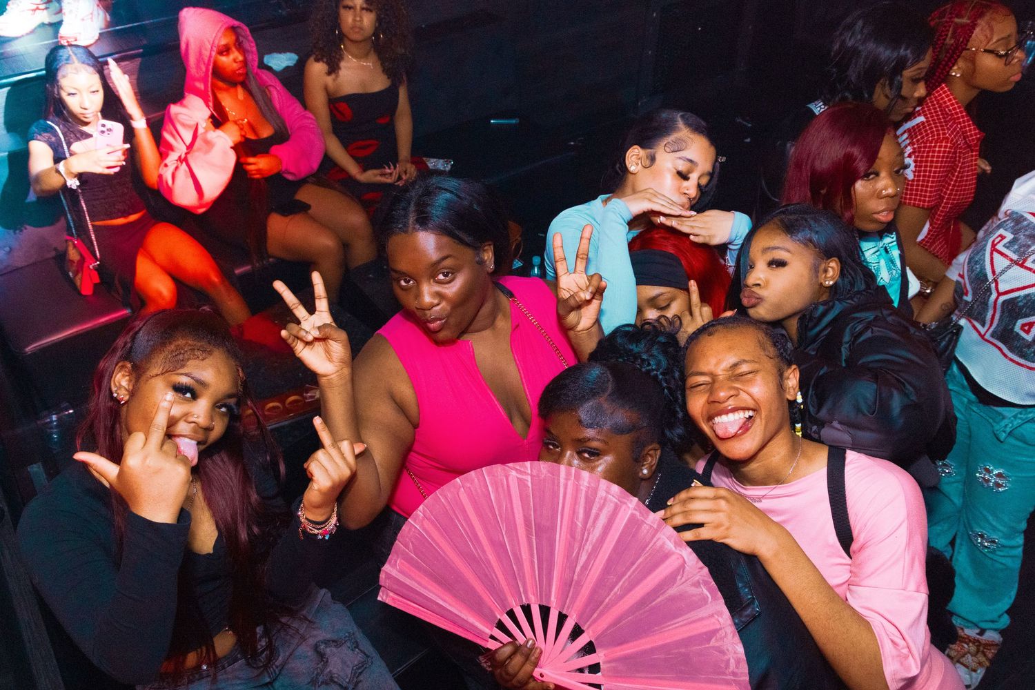 Group of young women posing and smiling at a nightclub party, one holding a pink decorative fan.