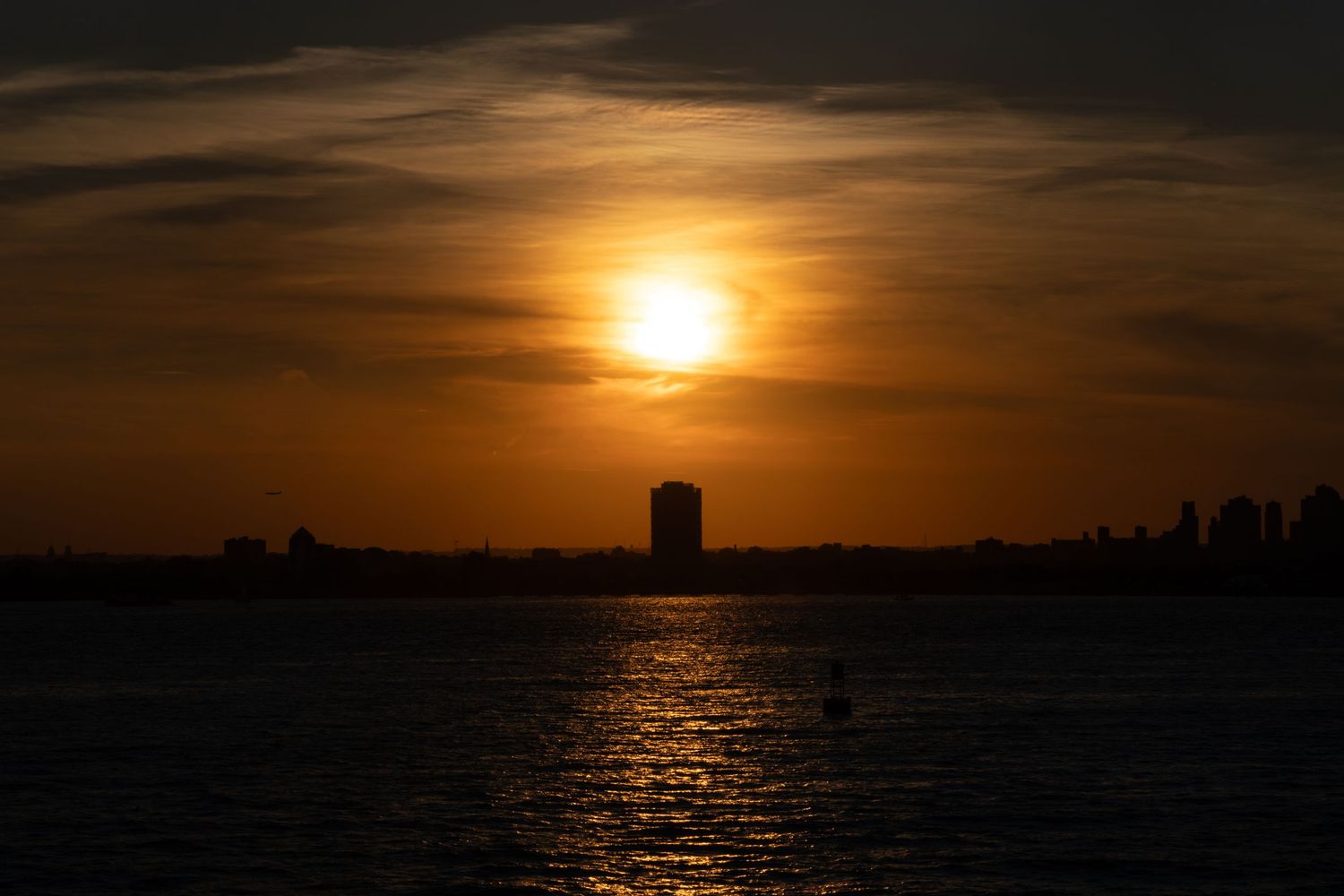 Golden sunset over calm water with a silhouetted skyline and tall building reflecting warm orange hues across the horizon.