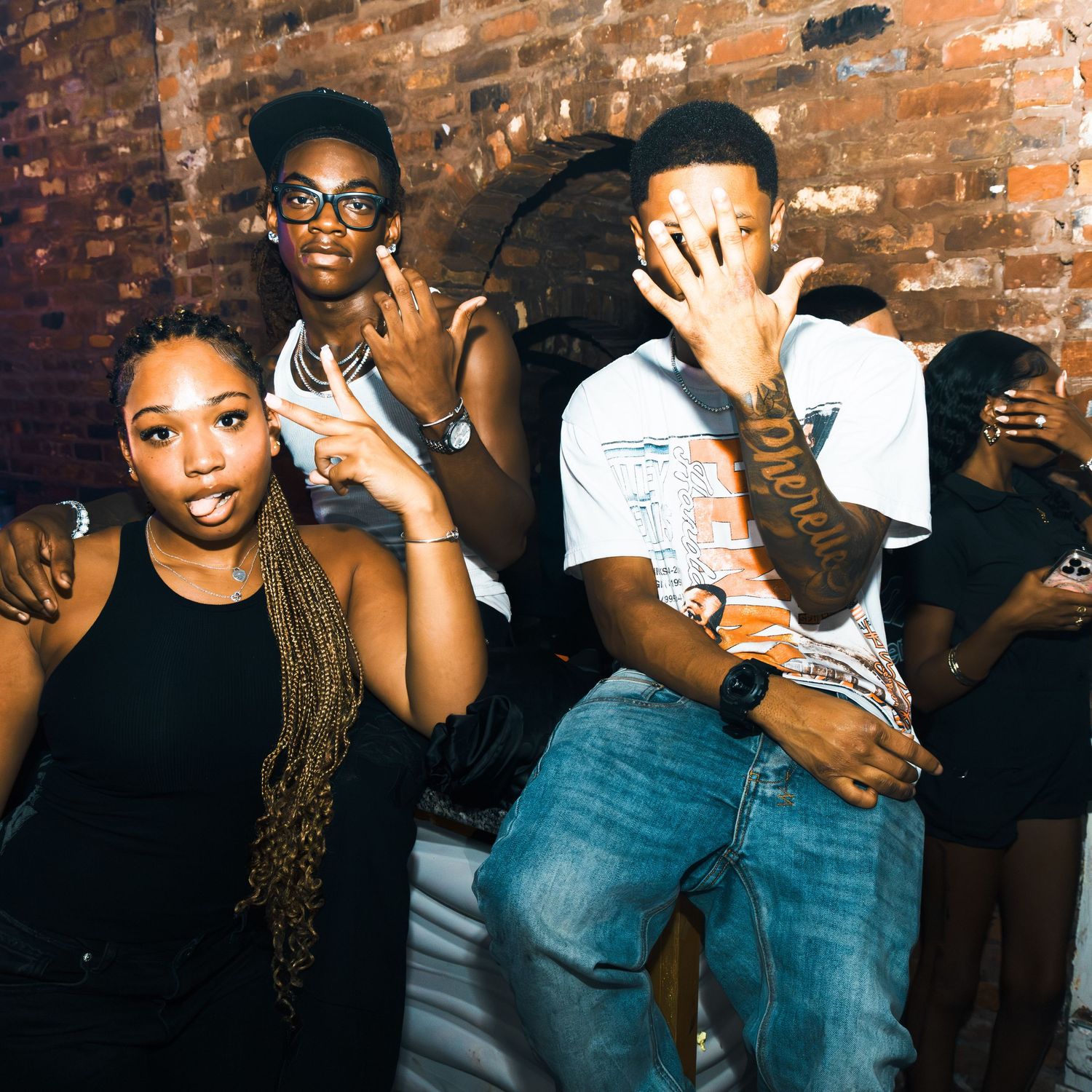 Group of young people posing and making hand gestures at a party near a brick wall.