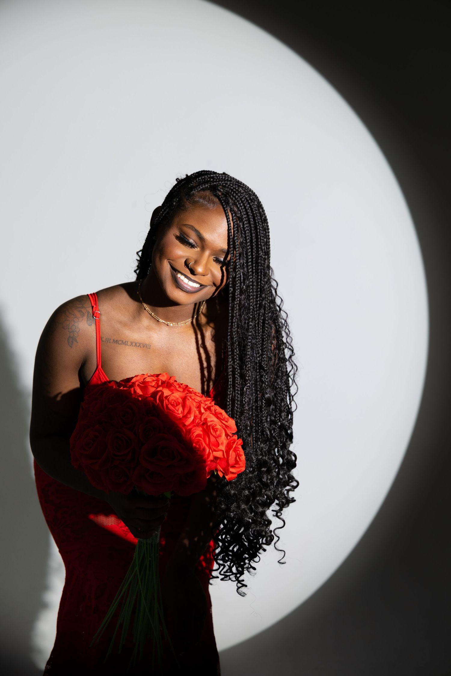 Smiling woman in red dress holding bouquet of red roses, with long braided hair, posed against white background.