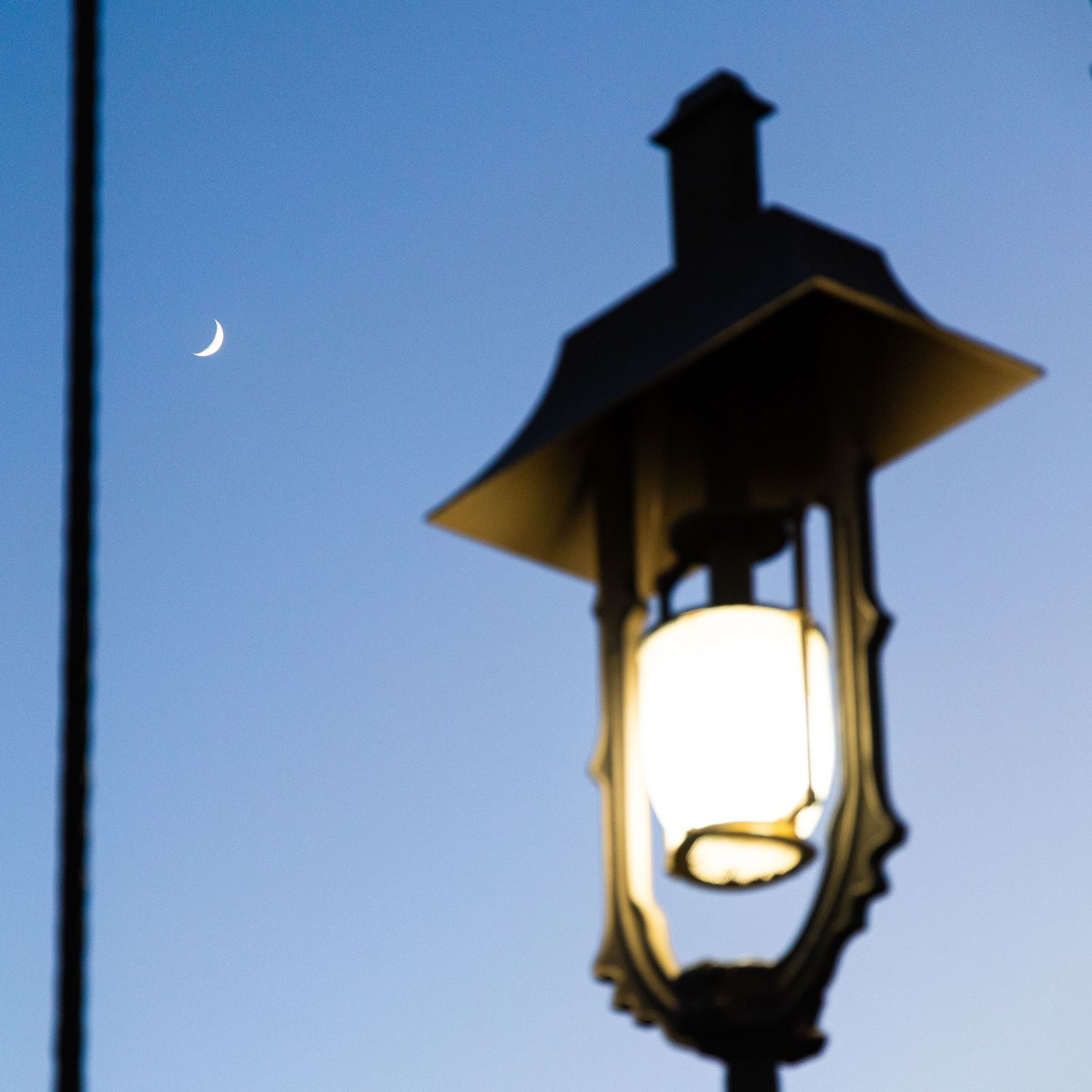 Ornate black street lamp glowing warmly against a twilight blue sky with a crescent moon visible in the background.