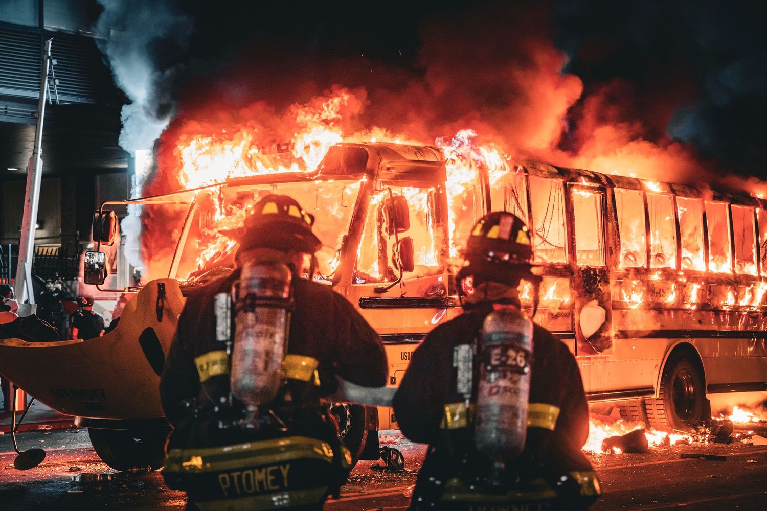 Two firefighters in gear stand before a fully engulfed burning bus at night, flames lighting the dark sky.