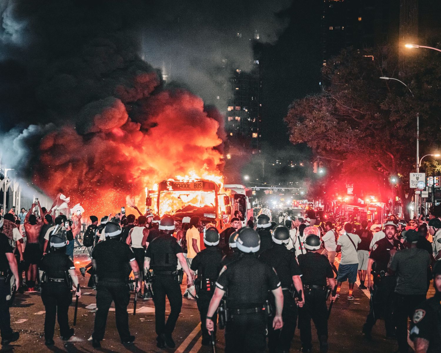 Police officers in riot gear face a large crowd amid massive red flames and smoke at a nighttime urban protest.