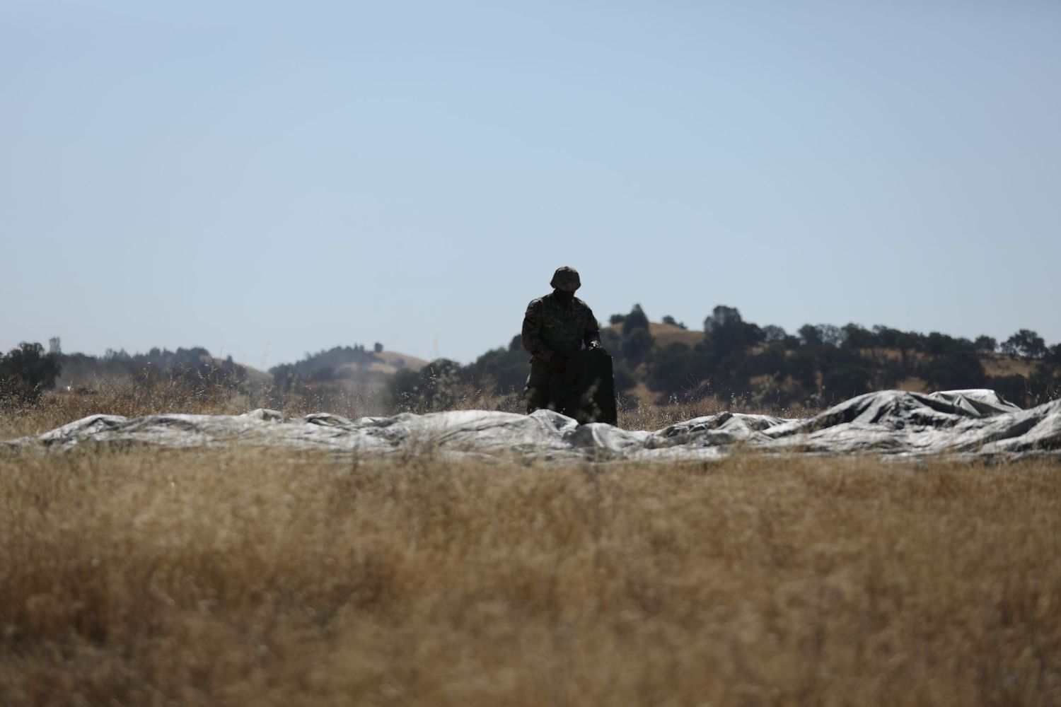 Lone military soldier silhouetted against a bright sky standing in a golden field beside white parachute fabric.