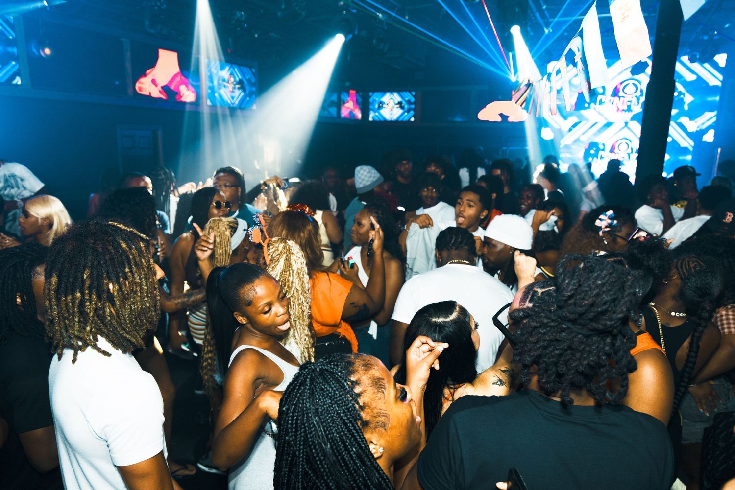 Crowded nightclub party with people dancing under bright stage lights and colorful visuals.