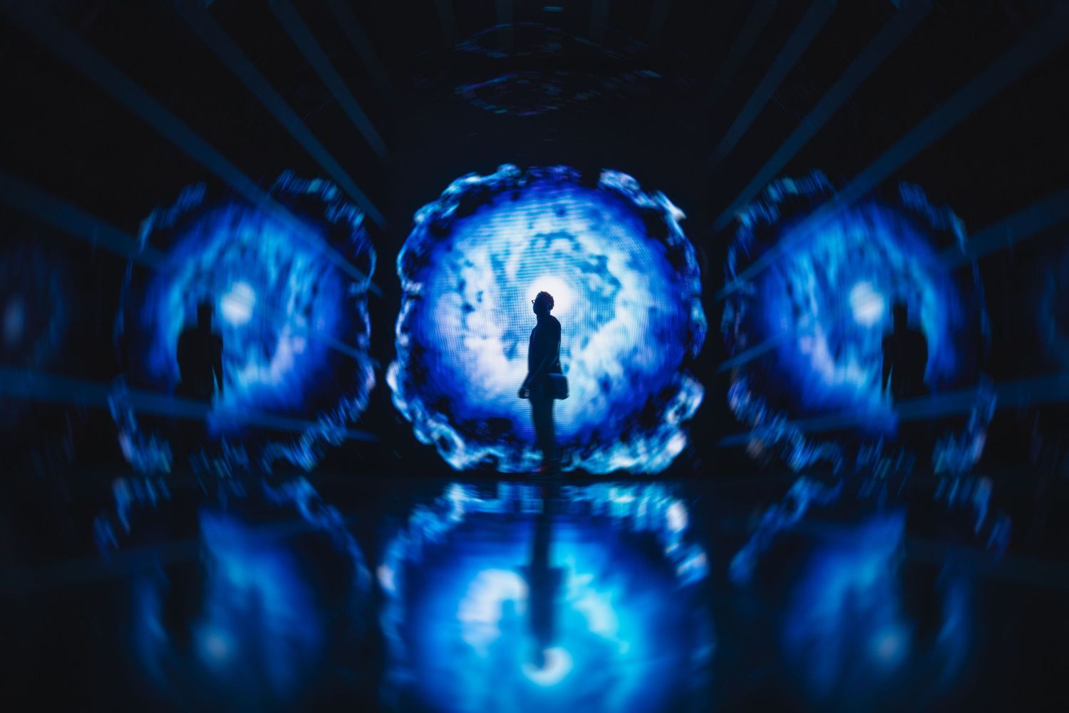 A silhouetted person stands before glowing blue circular portals in a dark, reflective immersive art installation.