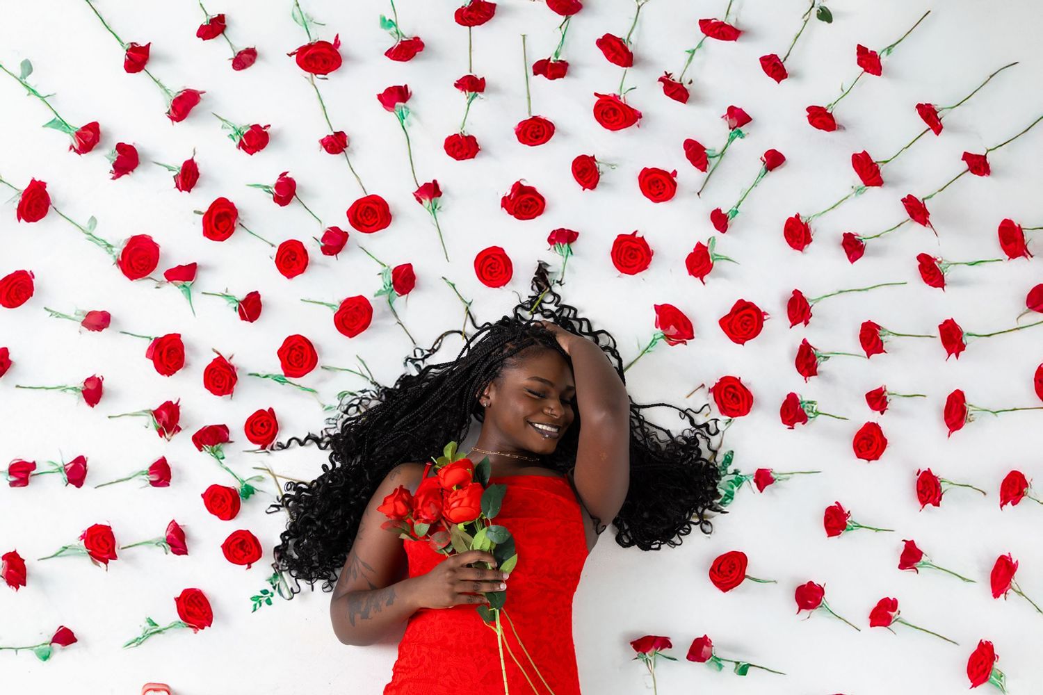 Woman in red dress lying among red carnations on white background, smiling and holding a bouquet of roses.
