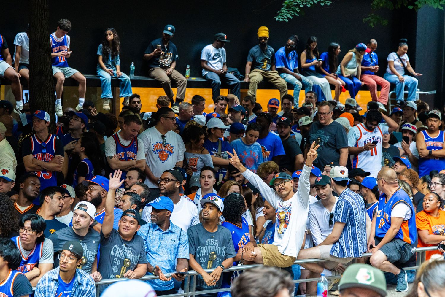 Excited New York Knicks fans cheering in stands, wearing blue and orange team gear at an outdoor NBA fan event.
