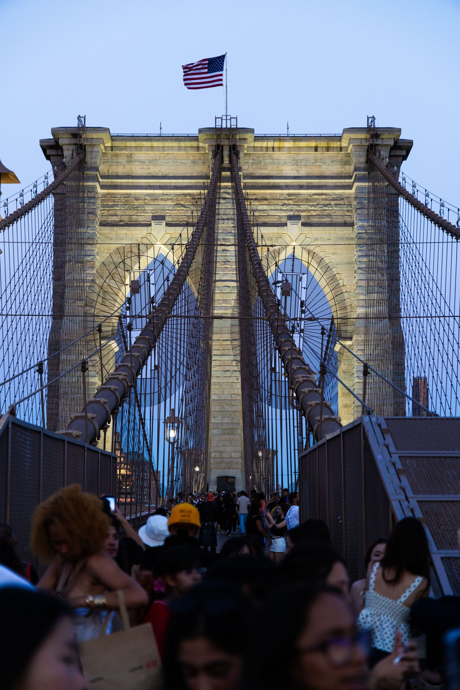 Crowds walk across the iconic Brooklyn Bridge at dusk with an American flag flying atop the historic stone tower.