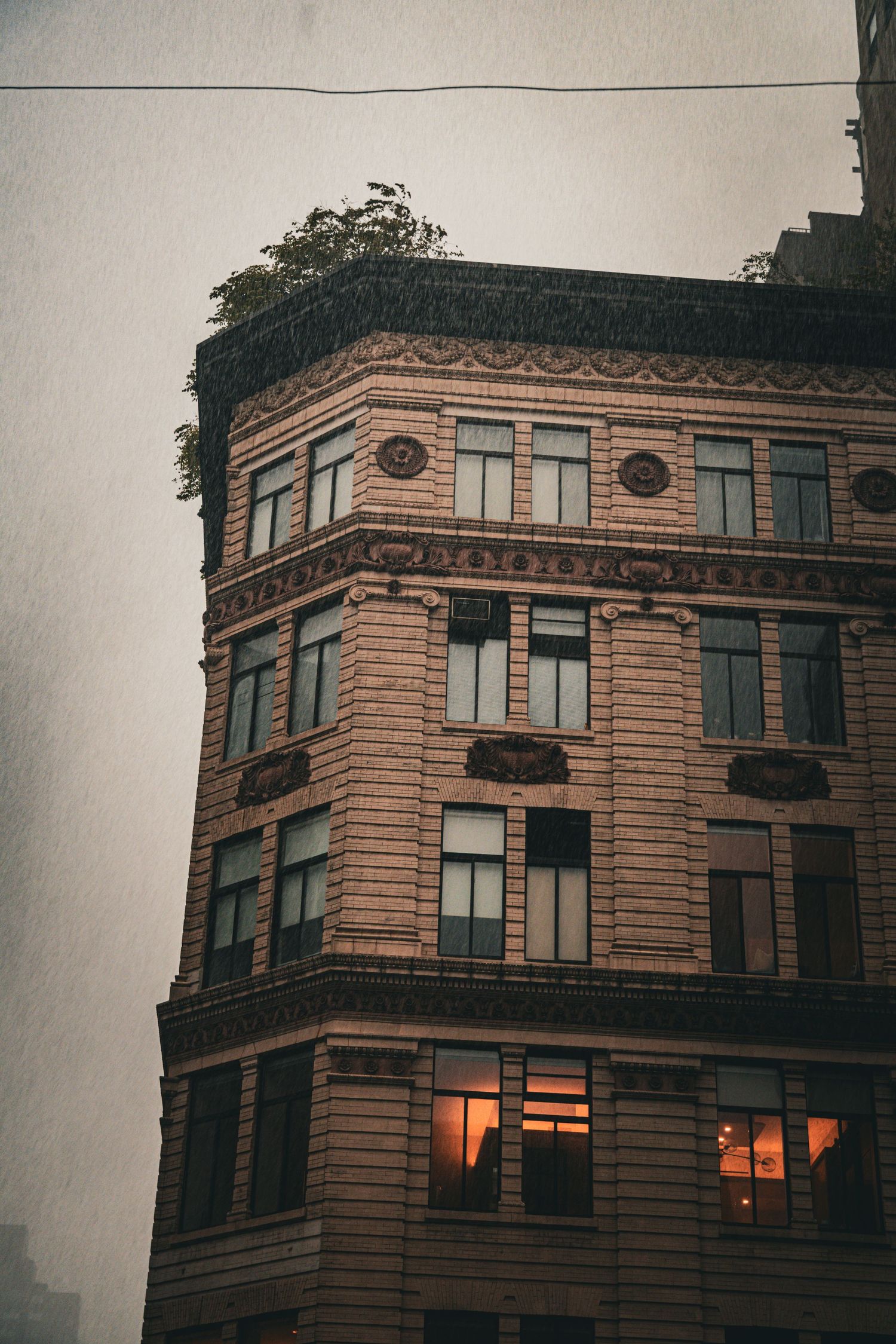 Historic brick building corner at dusk with ornate facade, glowing windows, and rooftop greenery under overcast sky.