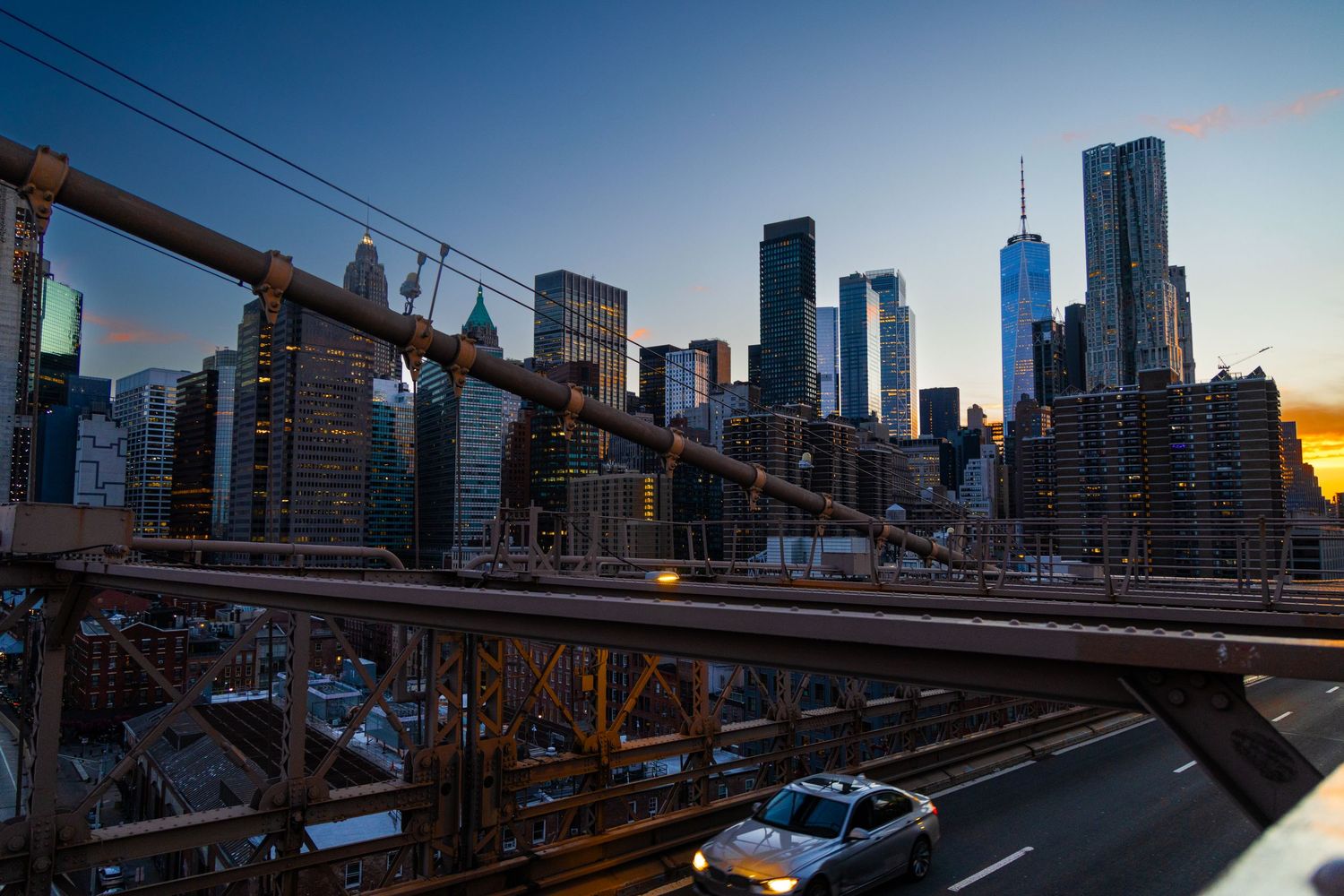 Brooklyn Bridge at dusk with Manhattan skyline and One World Trade Center glowing against a twilight sky.