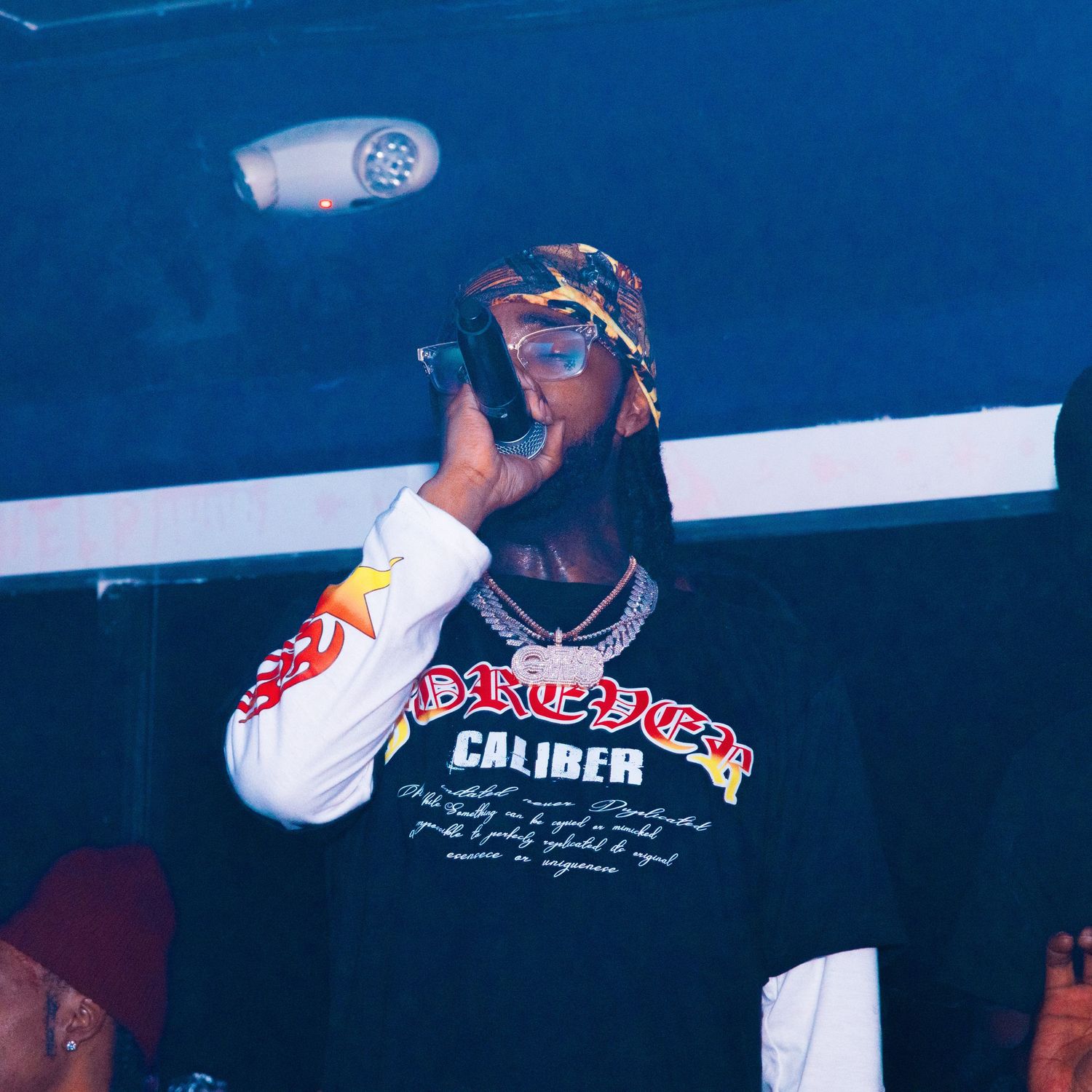 Rapper performing on stage holding microphone wearing Forever Caliber graphic tee and colorful headwrap under blue lights.