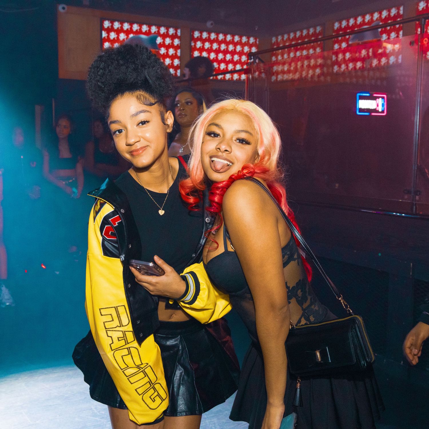 Two women pose together at a nightclub, one in a yellow jacket and one with pink hair in a black outfit.