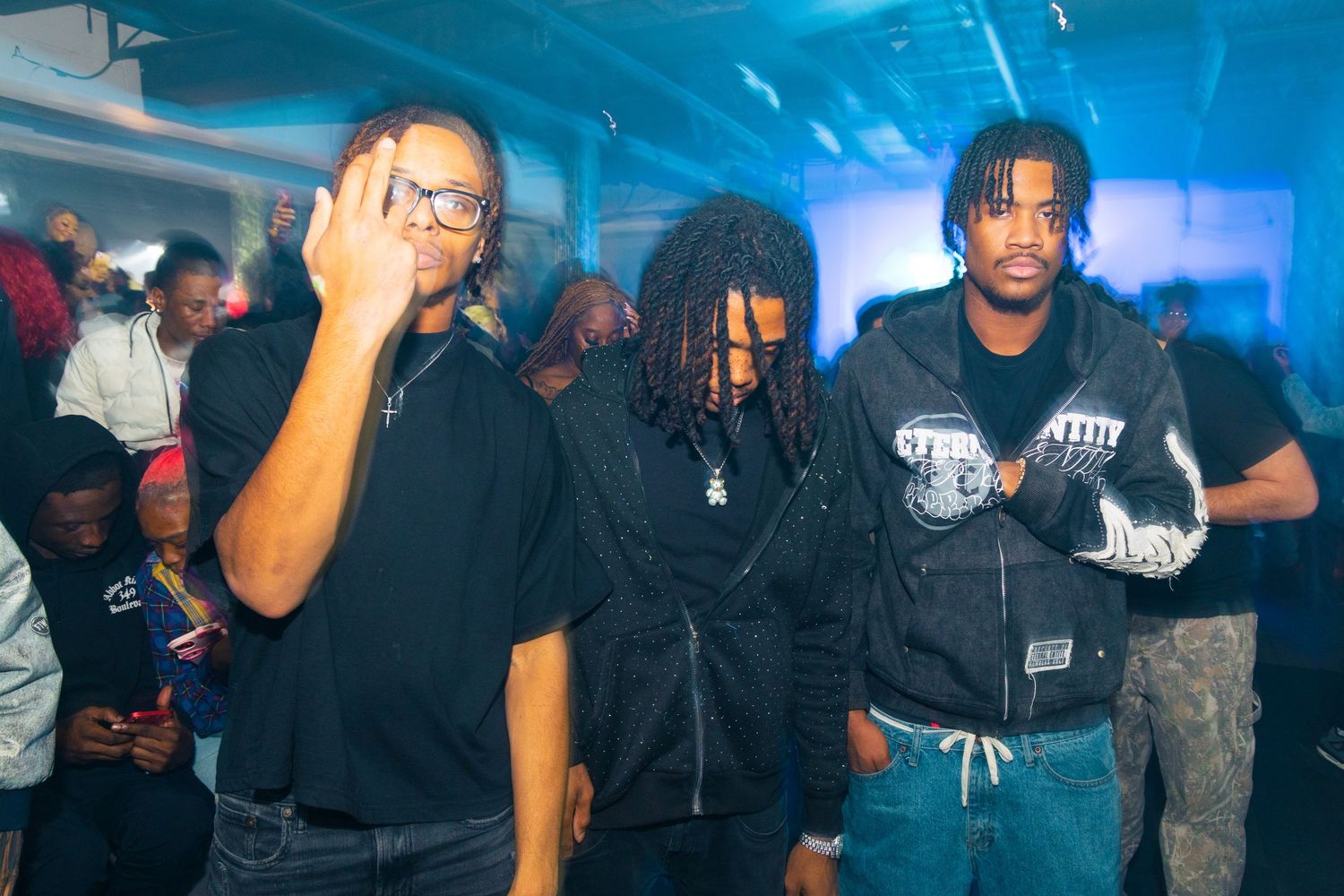 Three young men in black streetwear pose together at a crowded indoor event with blue ambient lighting.